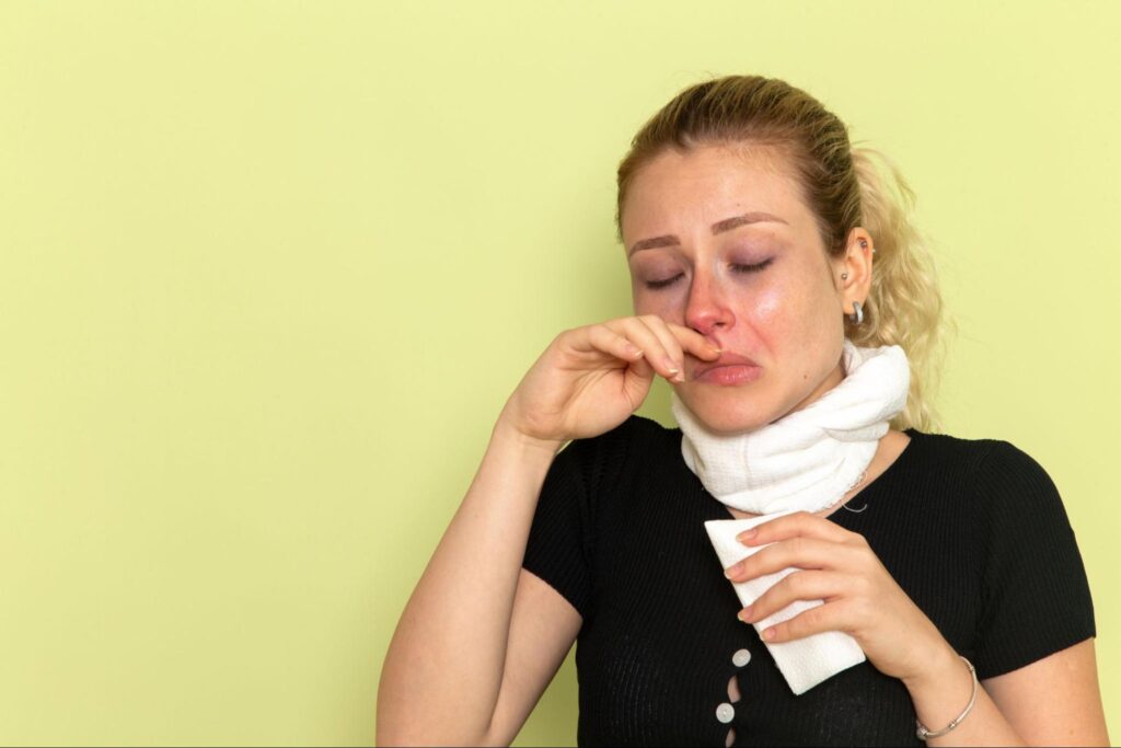 Woman with a white scarf around her neck holding a tissue and pinching her nose while dealing with allergy symptoms